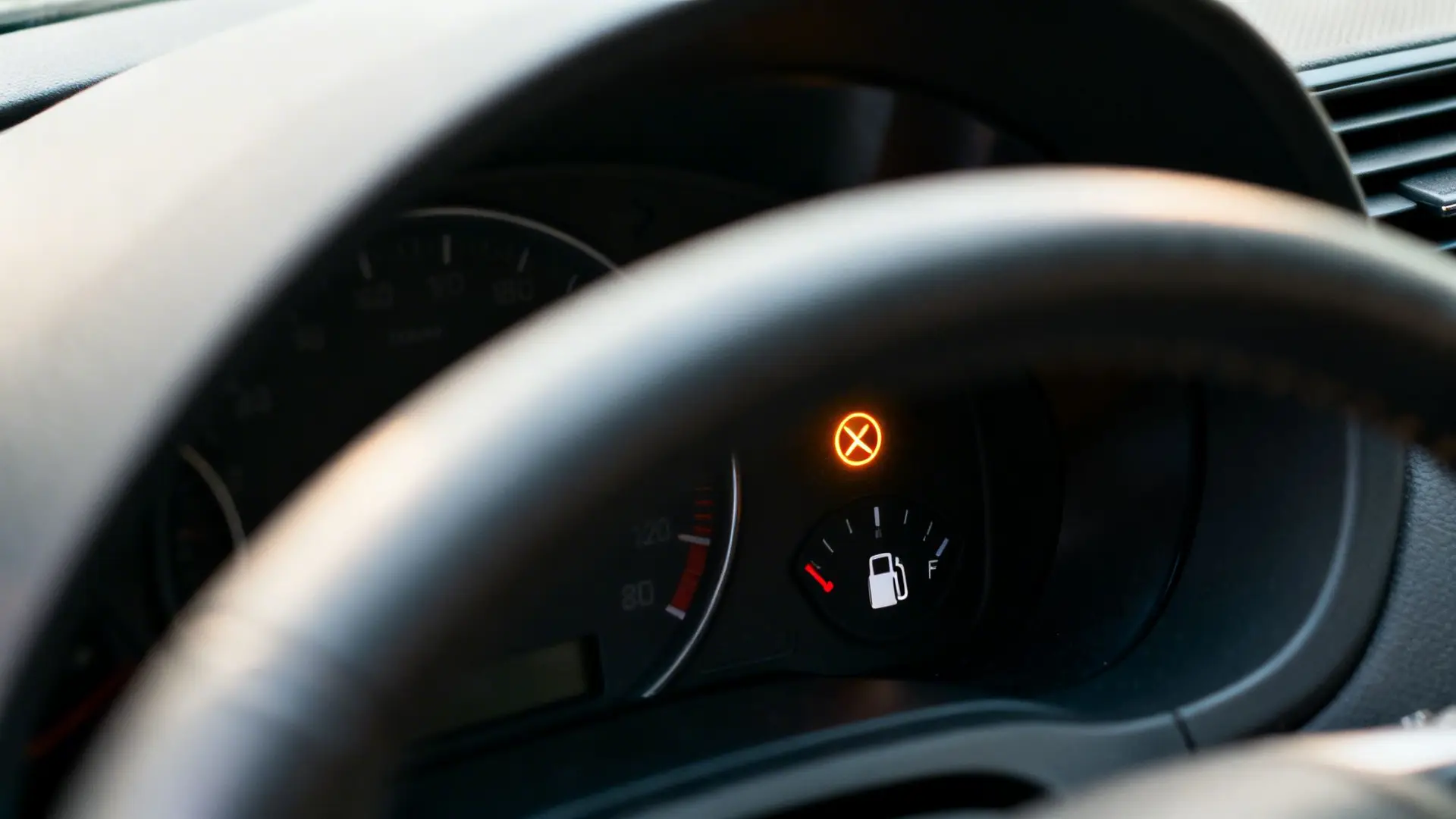 A close-up view of a car dashboard with the steering wheel partially covering it, showing a low fuel warning light and an orange warning symbol.