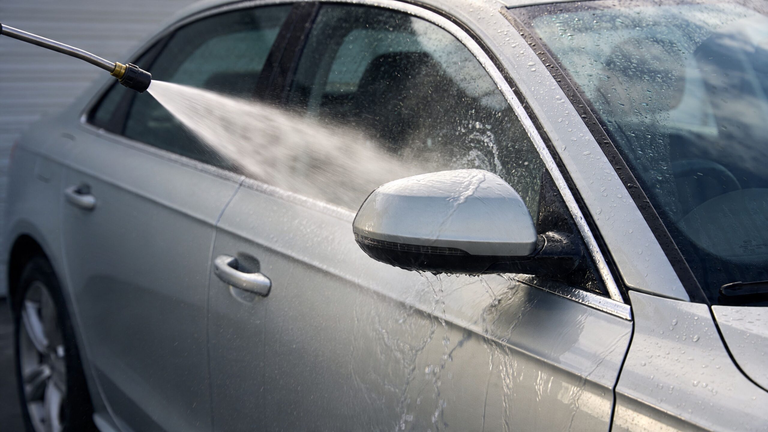 A pressure washer spraying water onto a silver car side mirror to clean the vehicle surface.