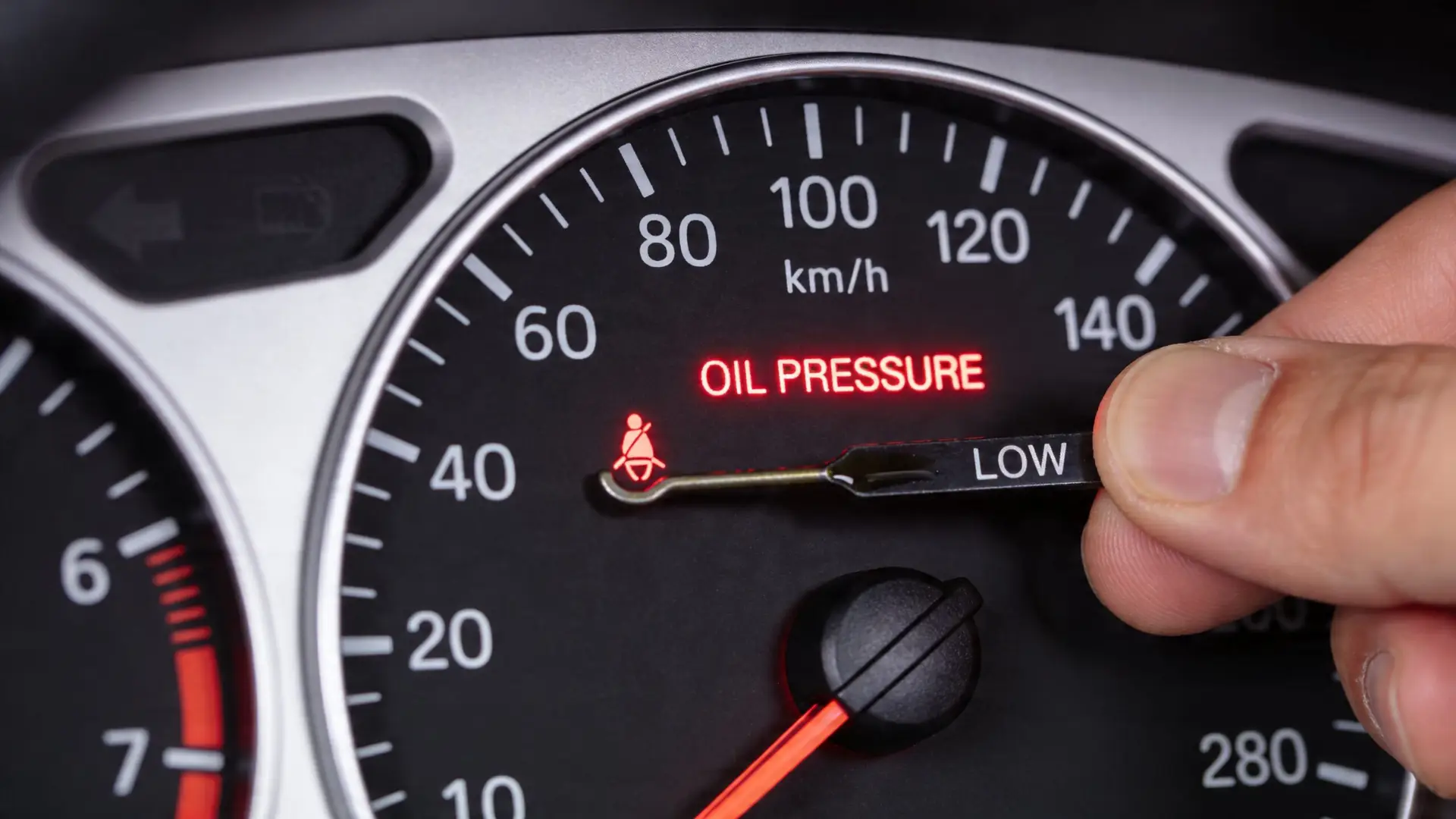 A hand holds a car oil dipstick showing low oil levels in front of a car dashboard
