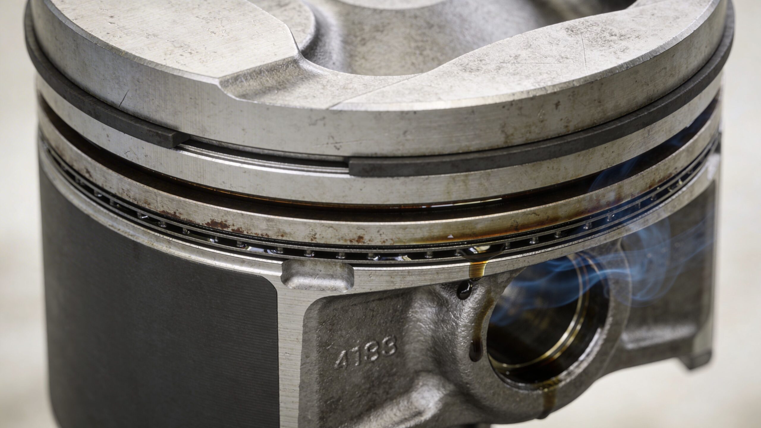 A close-up view of a metal engine piston with smoking oil dripping from its rings and body.