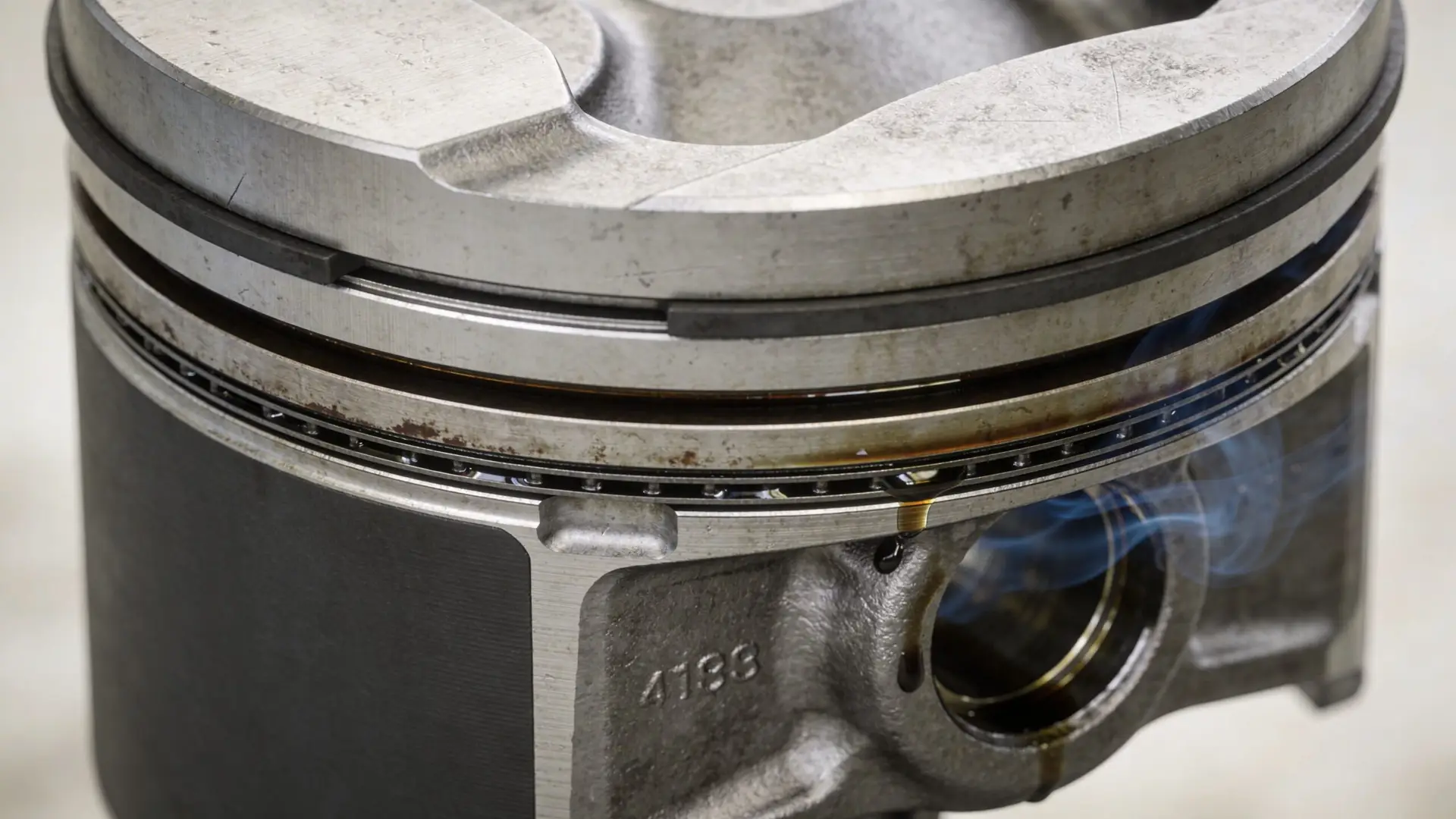 A close-up view of a metal engine piston with smoking oil dripping from its rings and body.