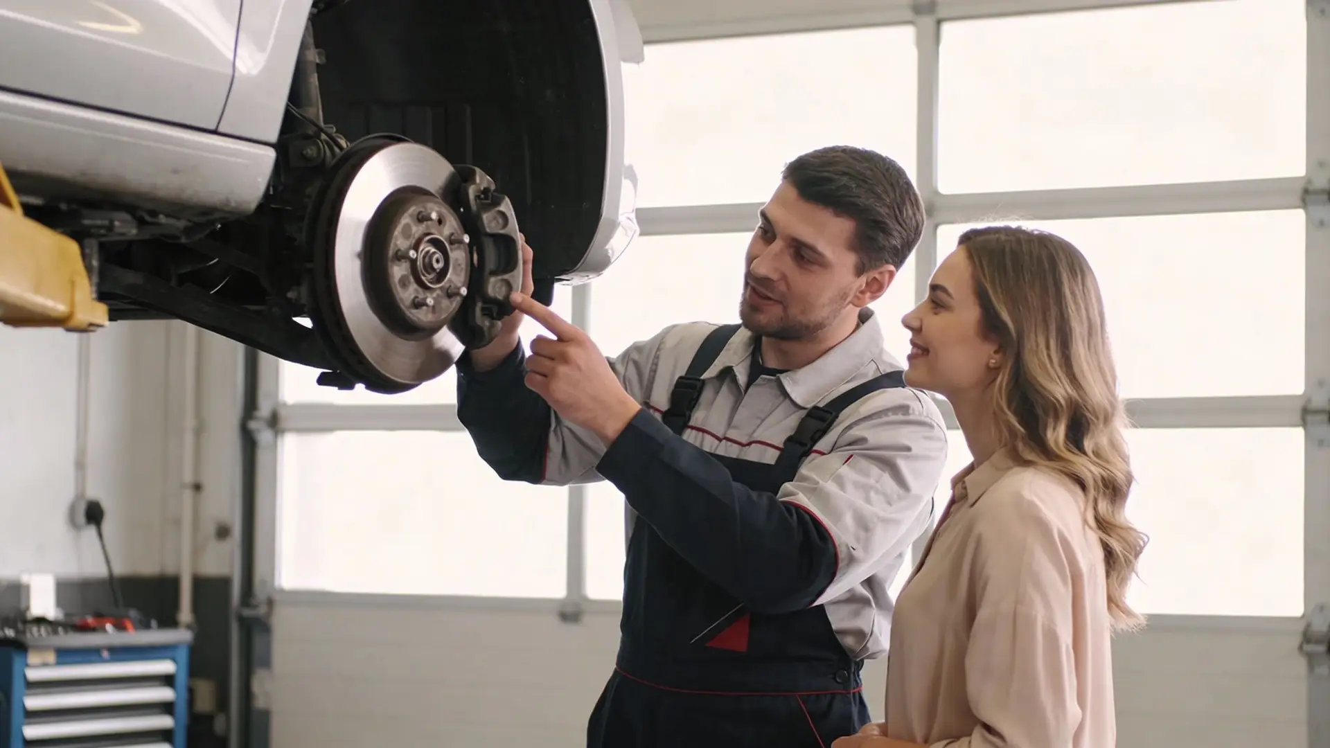 A professional mechanic explains car brake maintenance to a female customer in an auto repair shop.
